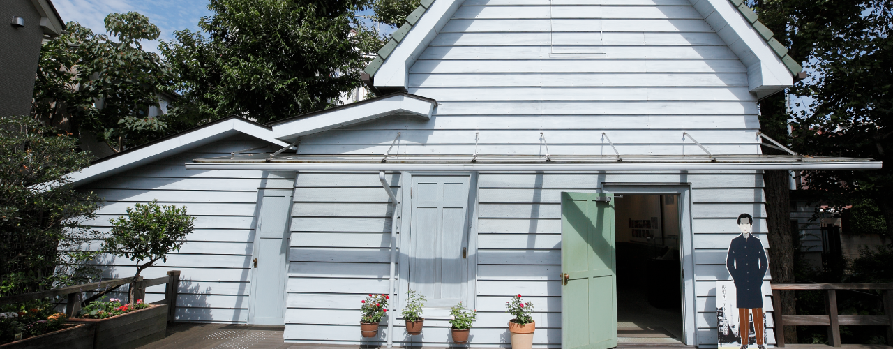 A pale blue wooden slated house with a cardboard cutout of Yuzo Saeki in front