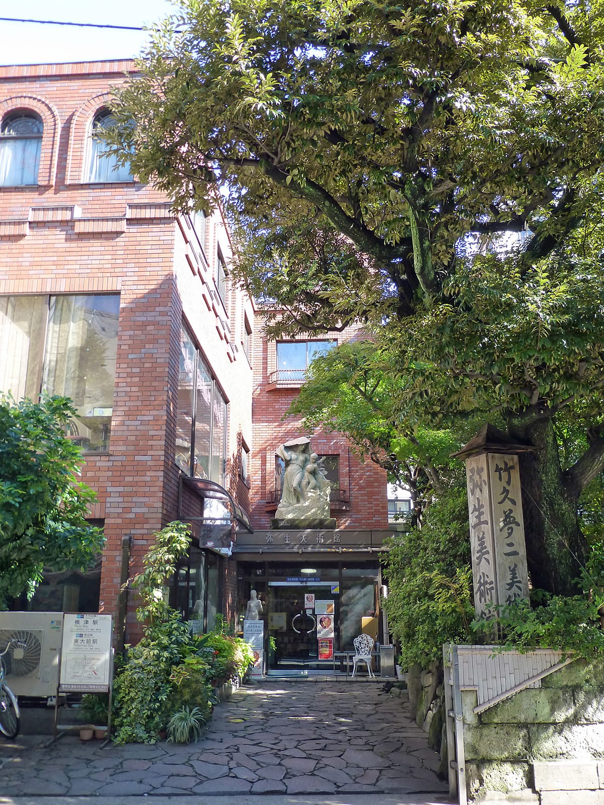 photo of the outside of a red brick building with a marble statue outside and a large statue over the entryway. A tree is in the foreground and a pilar bearing the name of the museum is next to it . 