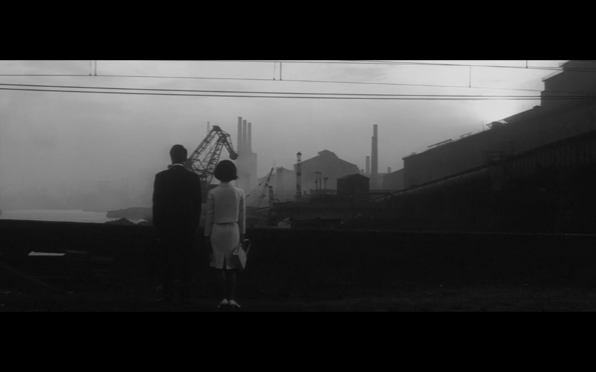 A man and a woman stand side by side, looking across the water at a construction site.