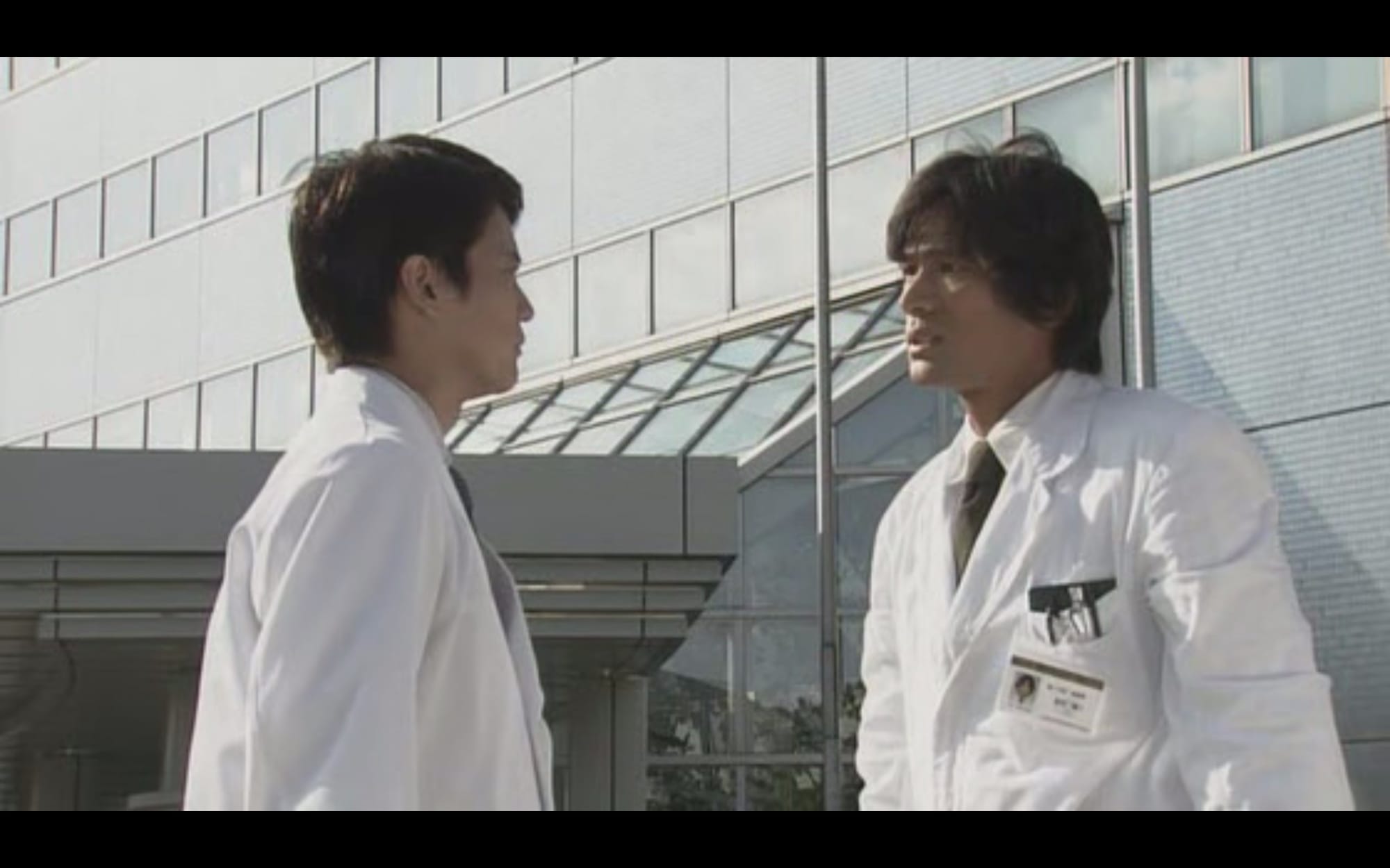 Two doctors stand facing each other in front of a hospital.